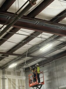 Crew member on a scissor lift cleaning exposed steel ceiling beams in a commercial warehouse