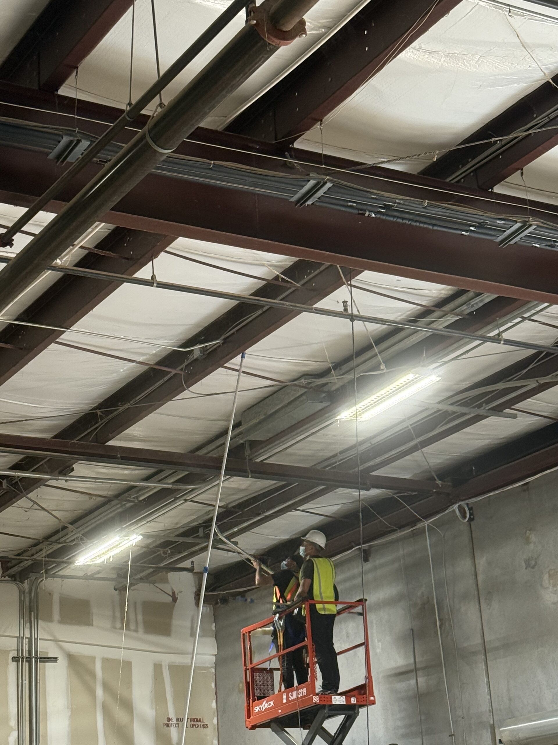Crew member on a scissor lift cleaning exposed steel ceiling beams in a commercial warehouse